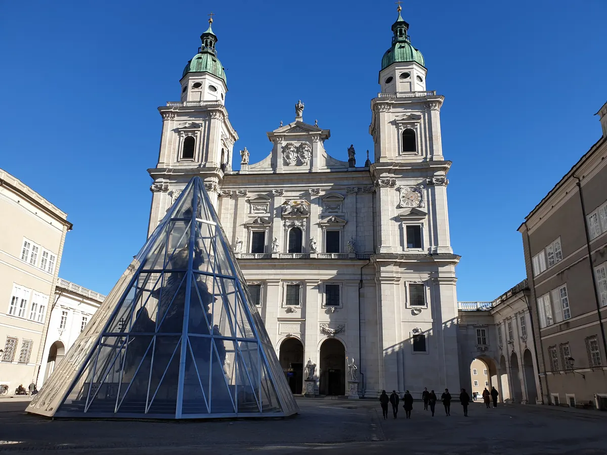 The west front of Salzburg Cathedral on Domplatz under a blue sky, with a glass pyramid covering the column in the square and the arcades at the right